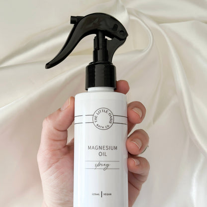 Hand holding a white spray bottle labeled 'Magnesium Oil' against a white fabric background